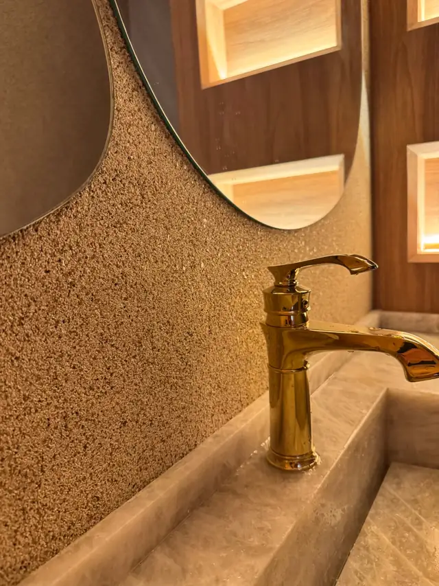 Lavabo com parede revestida em textura de pedras naturais, espelho orgânico e torneira dourada